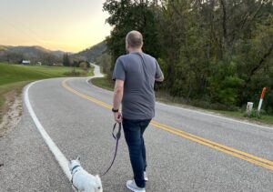 A man and his little white dog walking down a road that winds through the valley ahead of him. 