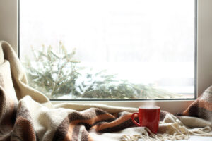 red mug of hot drink on a windowsill with a warm blanket, when behind is window is snow / cozy home atmosphere in the winter